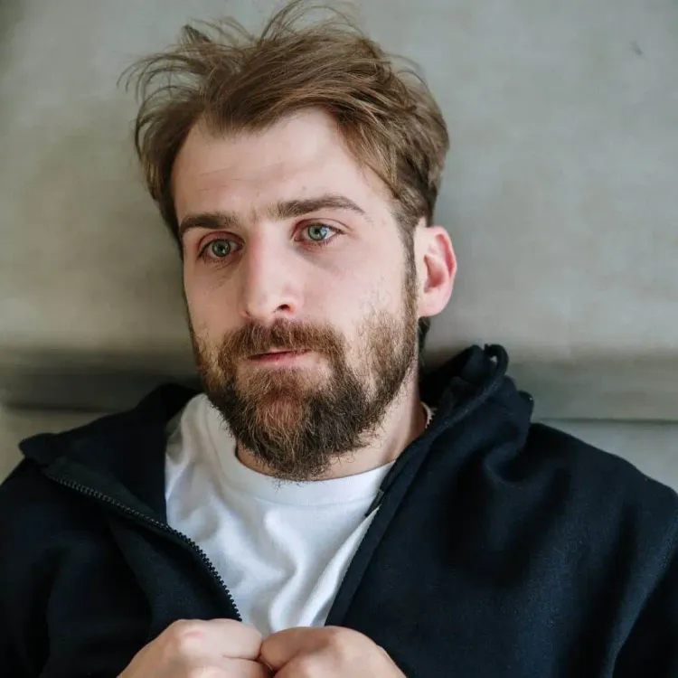 A bearded man with light brown hair, slightly messy, is lying on a gray surface. He is wearing a dark zip-up hoodie over a wh