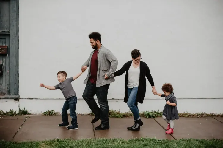 A family of four walks hand in hand on a sidewalk in front of a white wall. The father and mother are in the middle, holding