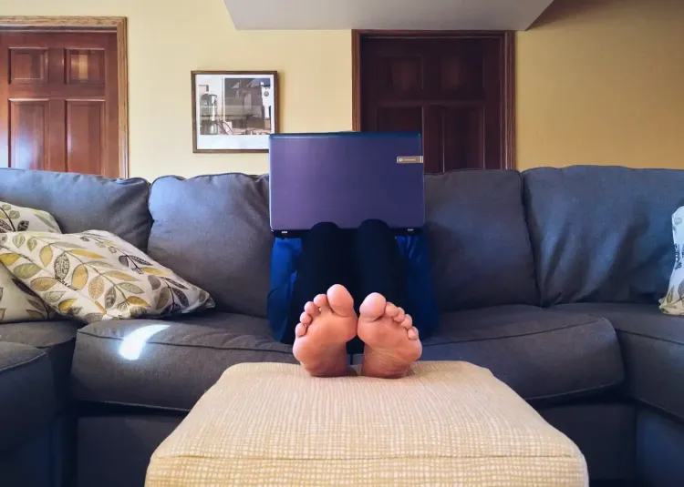 Person sitting on a gray couch with their feet up on a beige ottoman, using a laptop that hides their face. The background sh
