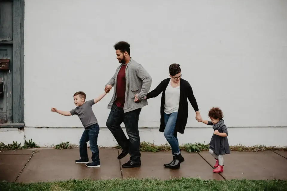 A family of four walks hand in hand on a sidewalk in front of a white wall. The father and mother are in the middle, holding